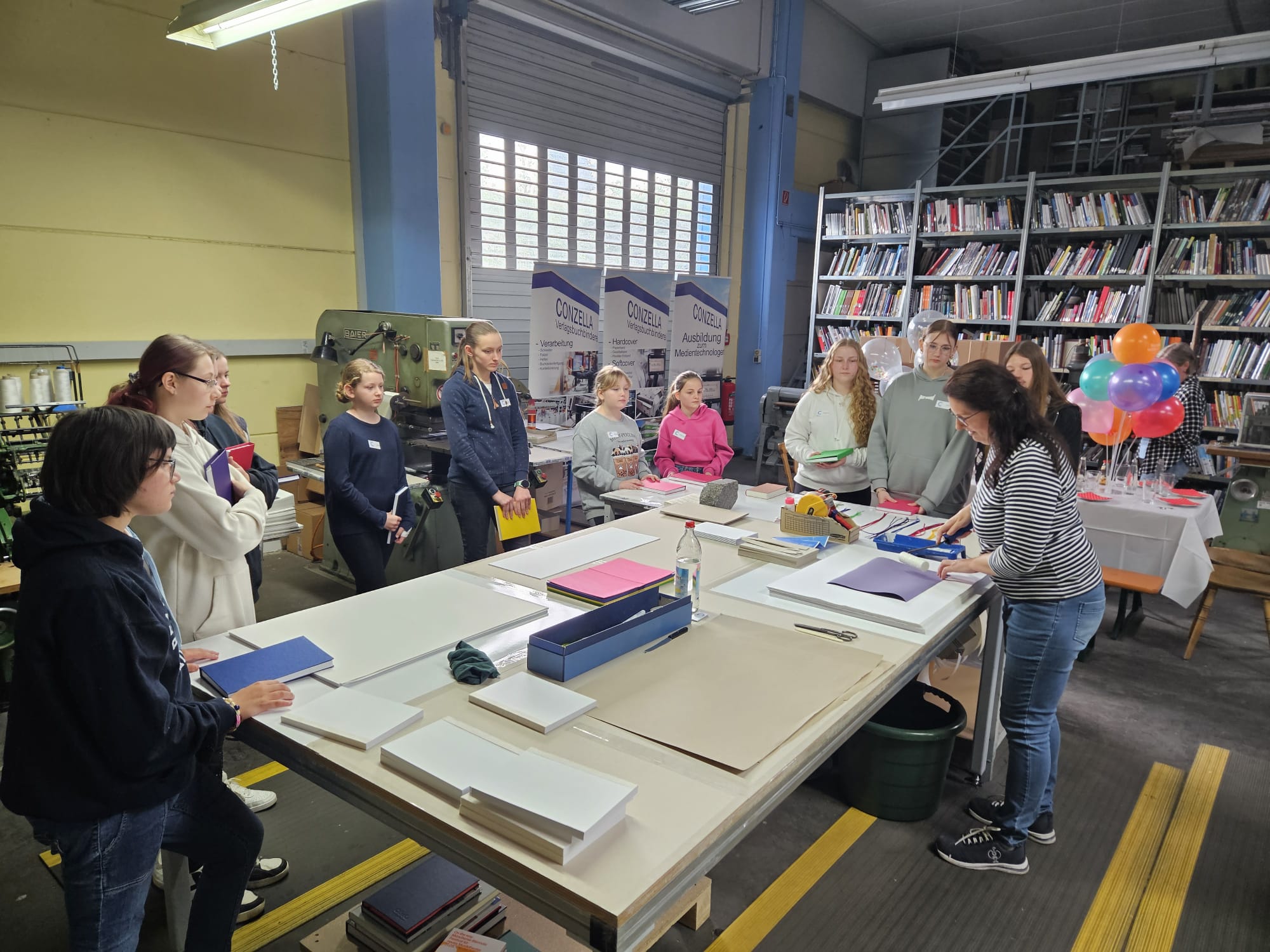 Bild vom Girls day 2024 bei der Conzella –Mit Leidenschaft zum Buch. Die Teilnehmer stehen in der Mustermacherei um einen Tisch und schauen zu, wie Buchdecken gemacht werden.