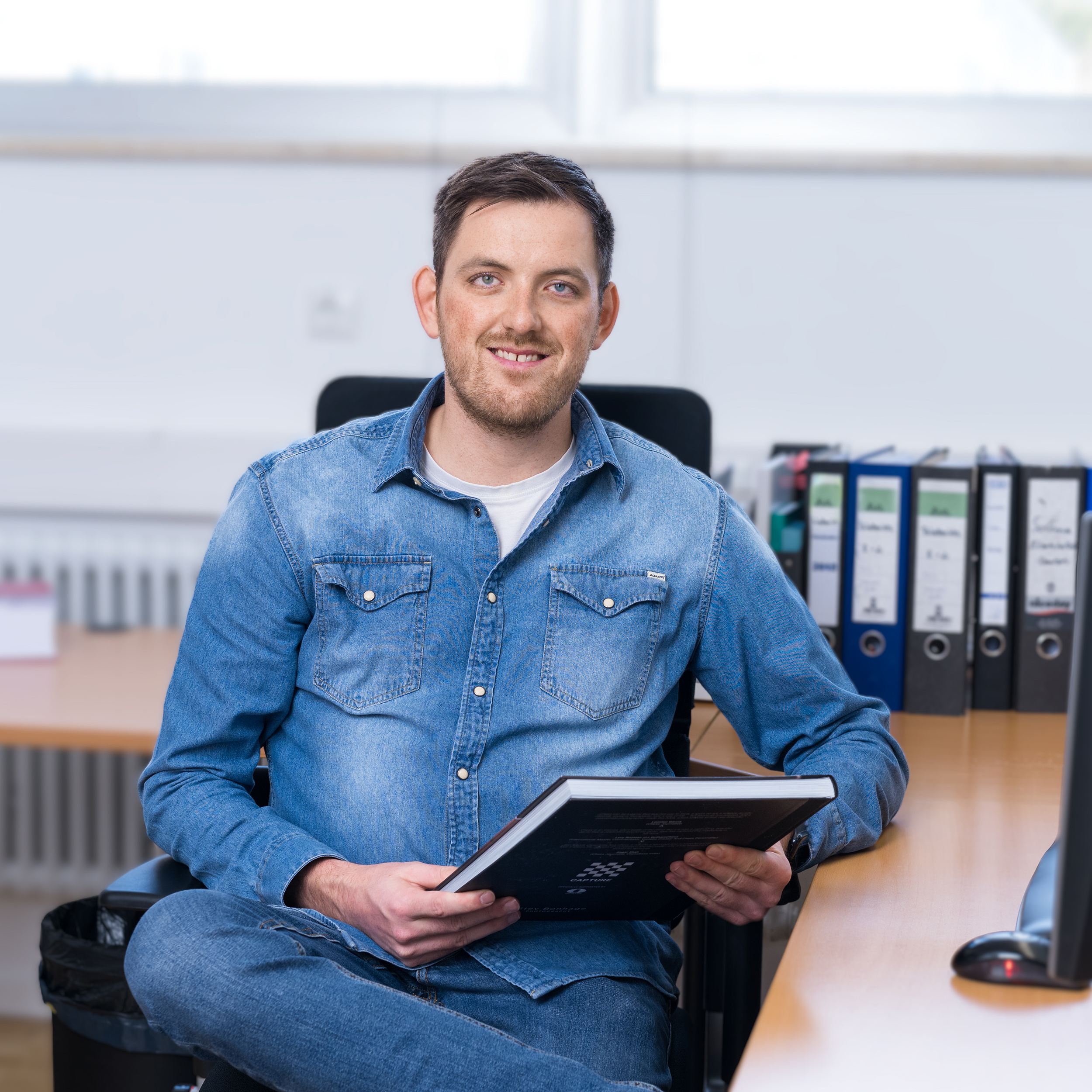 Porträt von Alois Osterholzer, Ausbilder und Assistent des Standortleiters bei der Conzella – Mit Leidenschaft zum Buch. Er trägt ein blaues Jeanshemd, sitzt an seinem Schreibtisch und hält ein Buch in den Händen. Im Vorder- und Hintergrund unscharf eine Büroszene mit Drucker, Computer, Ordnern und Telefon.