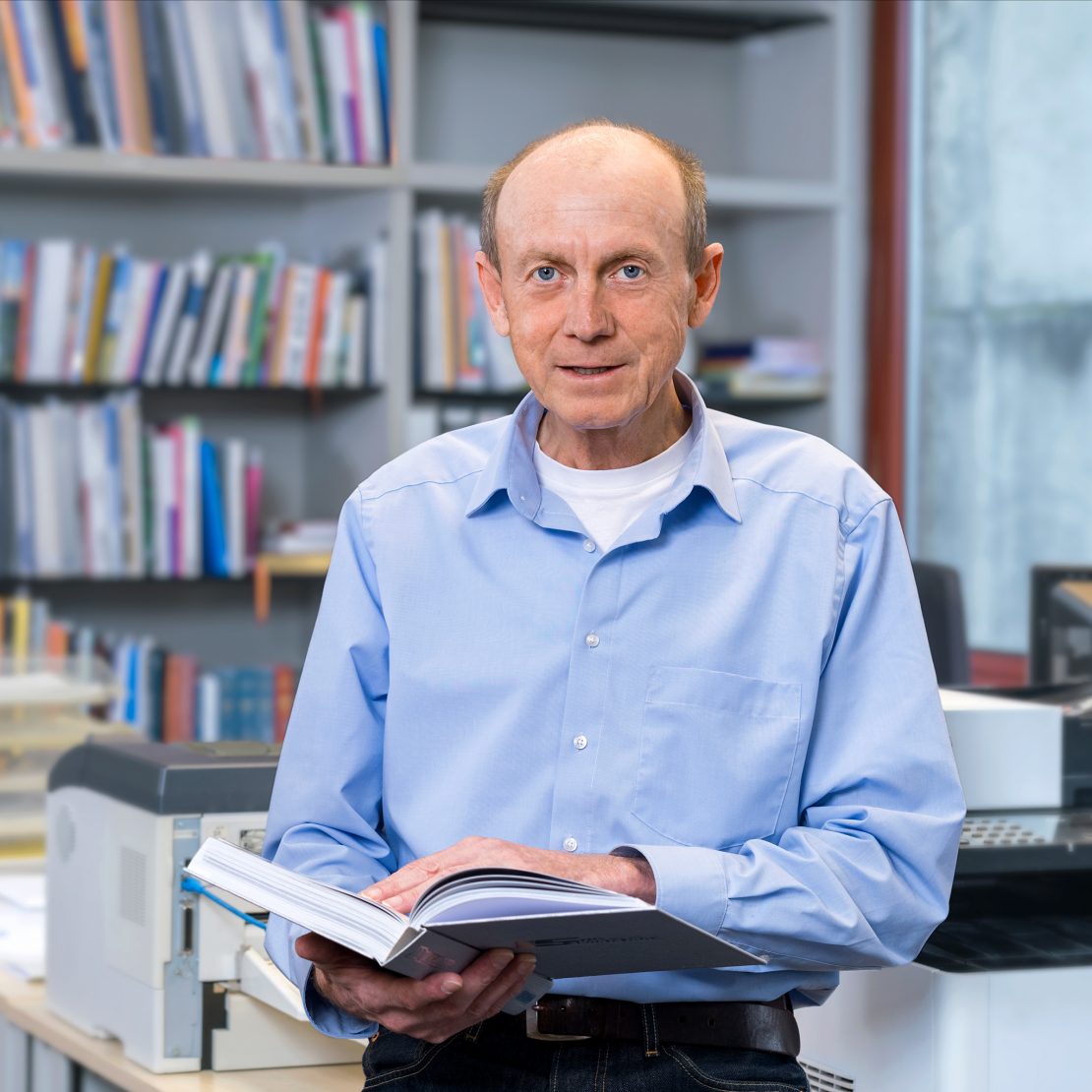 Porträt von Martin Wastian, Verkauf Innendienst bei der Conzella – Mit Leidenschaft zum Buch. Er trägt ein hellblaues Hemd und hält ein Buch in der Hand. Im Hintergrund unscharf eine Büroszene mit einem großen Bücherregal mit Büchern und Broschüren, die die Conzella produziert hat.